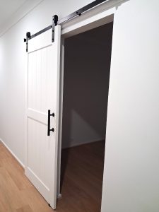 White sliding barn door with vertical paneling on a black metal track, partially open to a dark room.